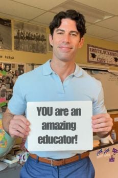 Male teacher in a classroom holding a sign that reads 'YOU are an amazing educator!'
