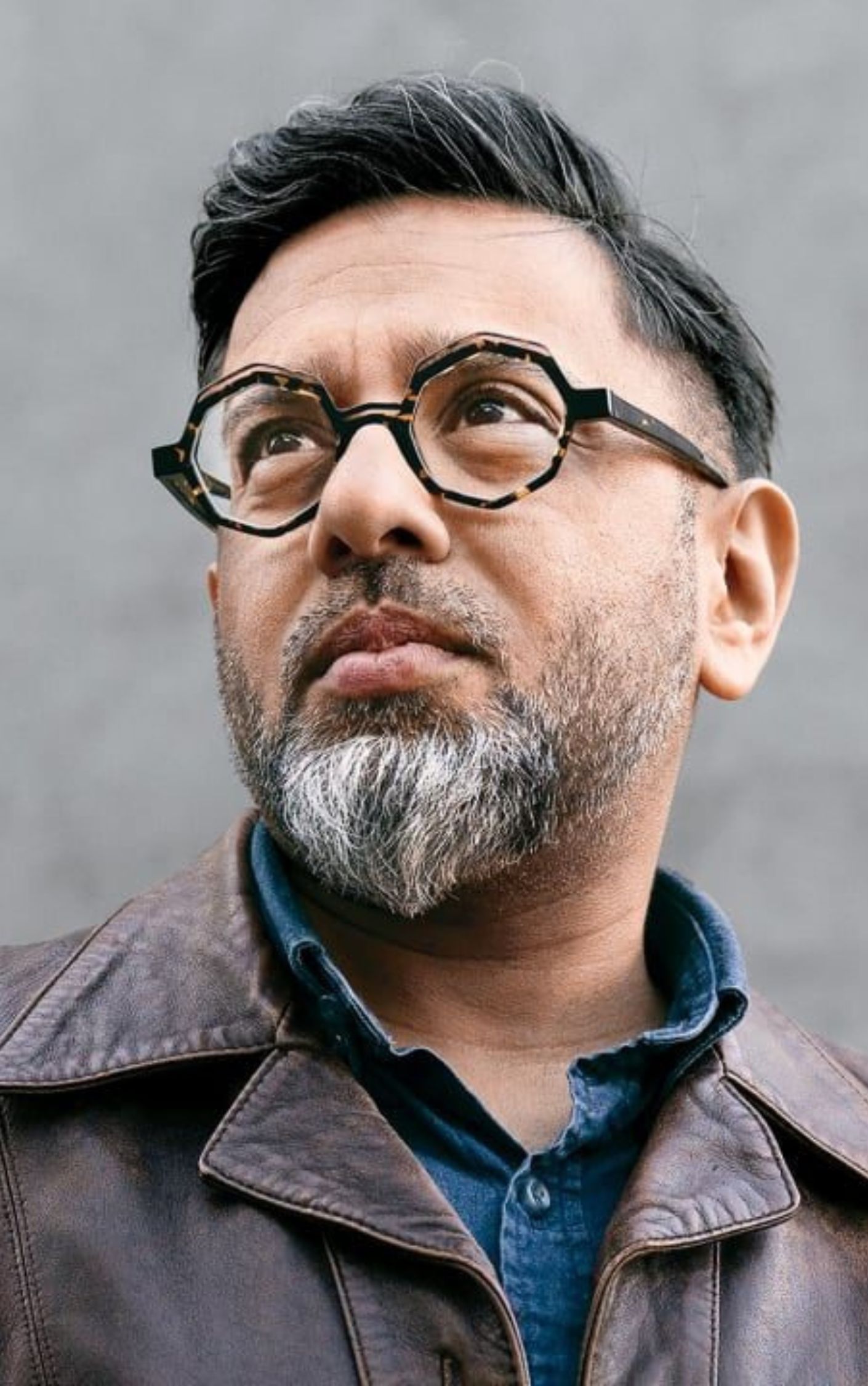 Portrait of Saket Soni, an Indian American labor leader, wearing a leather jacket, blue shirt, and round tortoiseshell glasses, looking thoughtfully into the distance.