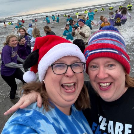 Two smiling co-teachers wearing festive hats take a selfie during a winter beach event, with students and participants splashing in the ocean behind them.